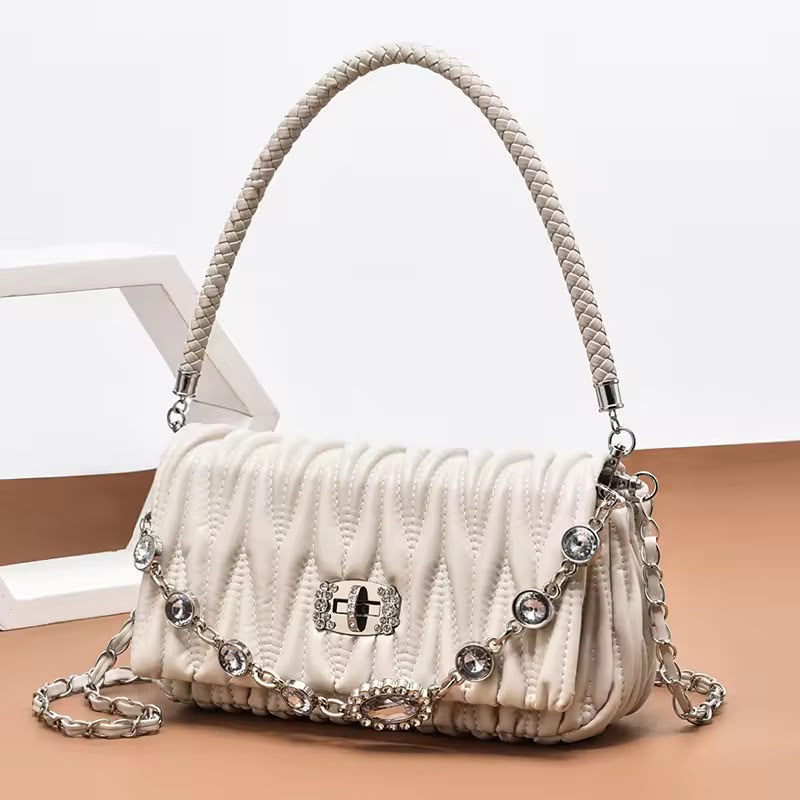 A cream-colored quilted shoulder bag with a braided top handle, silver-tone chain strap, and rhinestone embellishments, displayed on a tan surface against a white background.