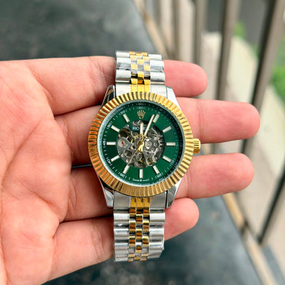 A two-tone stainless steel and gold wristwatch with a green dial is held in a person's hand. The watch features a fluted gold-tone bezel, a green dial with skeletonized details, and a matching two-tone metal link bracelet.