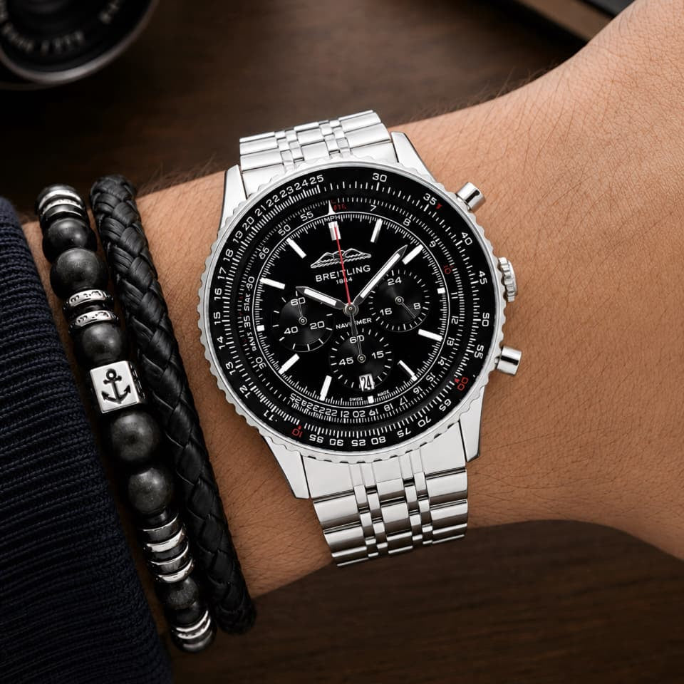 A silver stainless steel wristwatch with a black dial and tachymeter bezel, shown worn on a person's wrist alongside black beaded bracelets, set against a dark wooden desk background with blurred camera and notebook props.