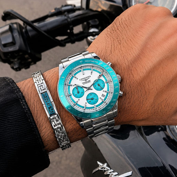A silver-toned metal wristwatch with a bright turquoise blue bezel and matching blue subdials, worn on a person's wrist next to a silver bracelet, with a motorcycle handlebar and tank visible in the background.